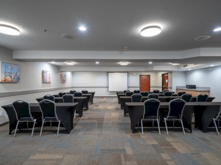 A conference room with multiple rows of tables and chairs, a projector screen at the front, framed artwork on the walls, and overhead lighting.