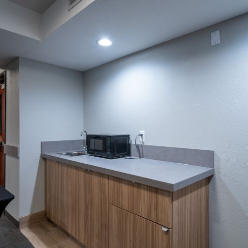 The image shows a simple kitchenette area with a microwave on the countertop, wooden cabinets below, and bright overhead lighting.