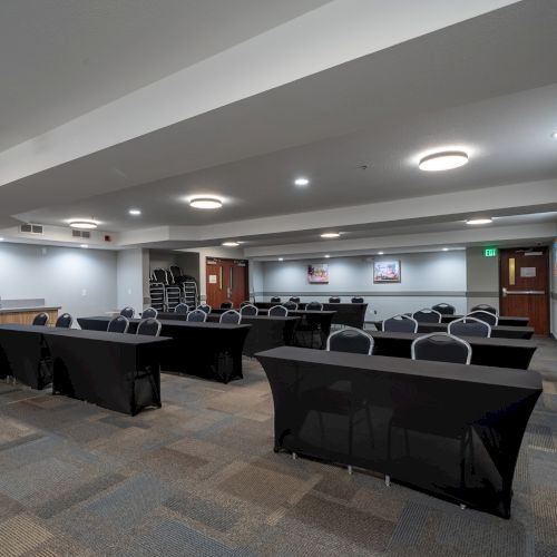 The image shows a conference room with black tablecloth-covered tables, chairs, a small kitchen area, and wall art. The room is well-lit by ceiling lights.
