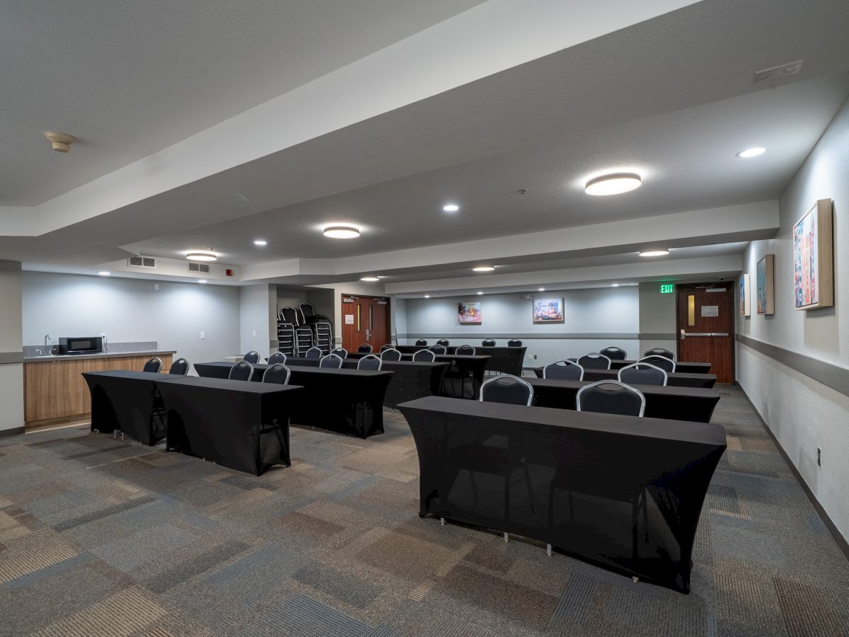 A conference room with multiple black-covered tables, chairs, a counter with a microwave, and light fixtures on the ceiling.